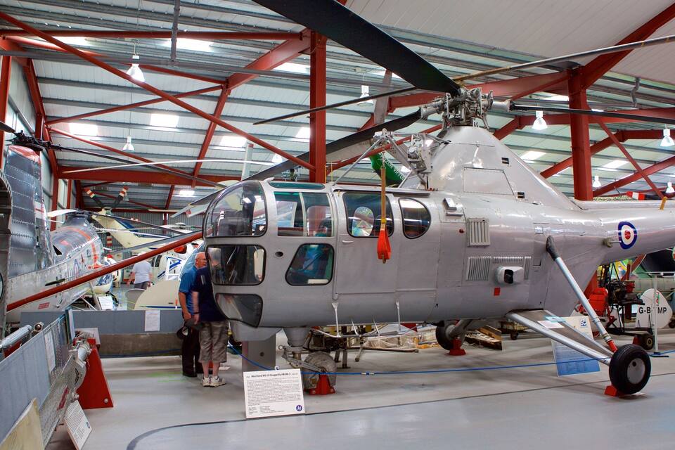 Un helicóptero militar dentro de un hangar con vigas rojas y otros aviones al fondo.