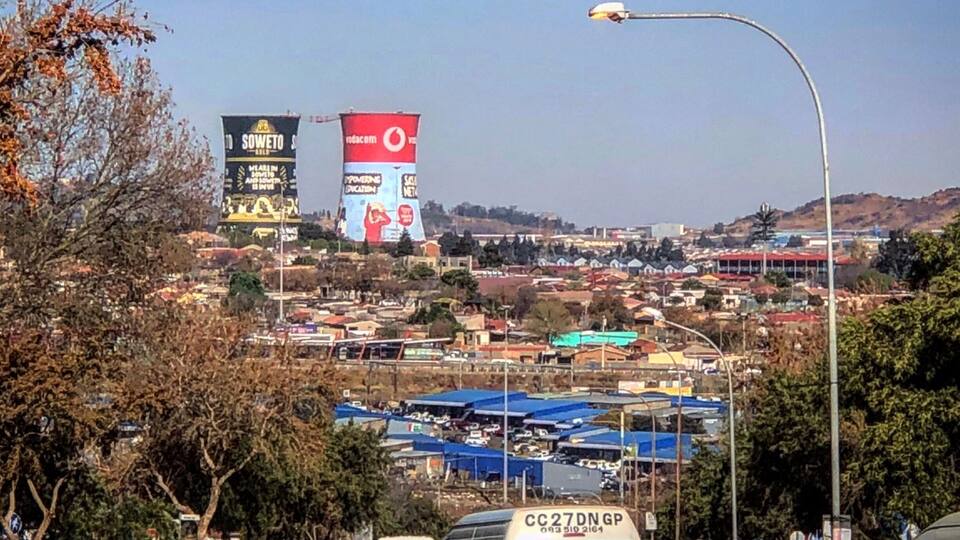 Paisagem urbana com um grande outdoor, uma rua com veículos e uma pessoa sentada em uma rocha.