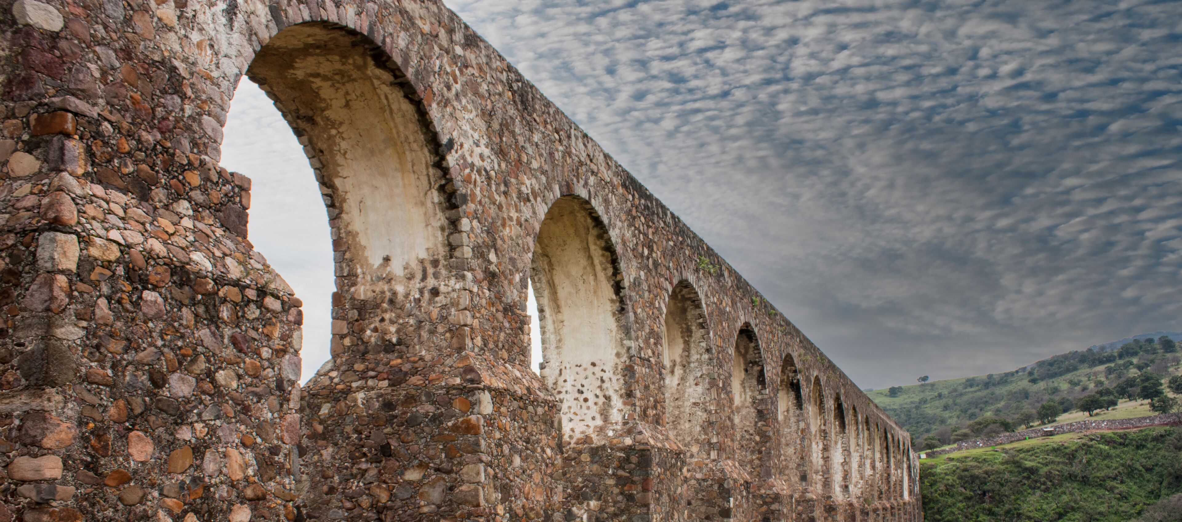 An ancient stone bridge with multiple arches, set against a cloudy sky and surrounded by green hills.