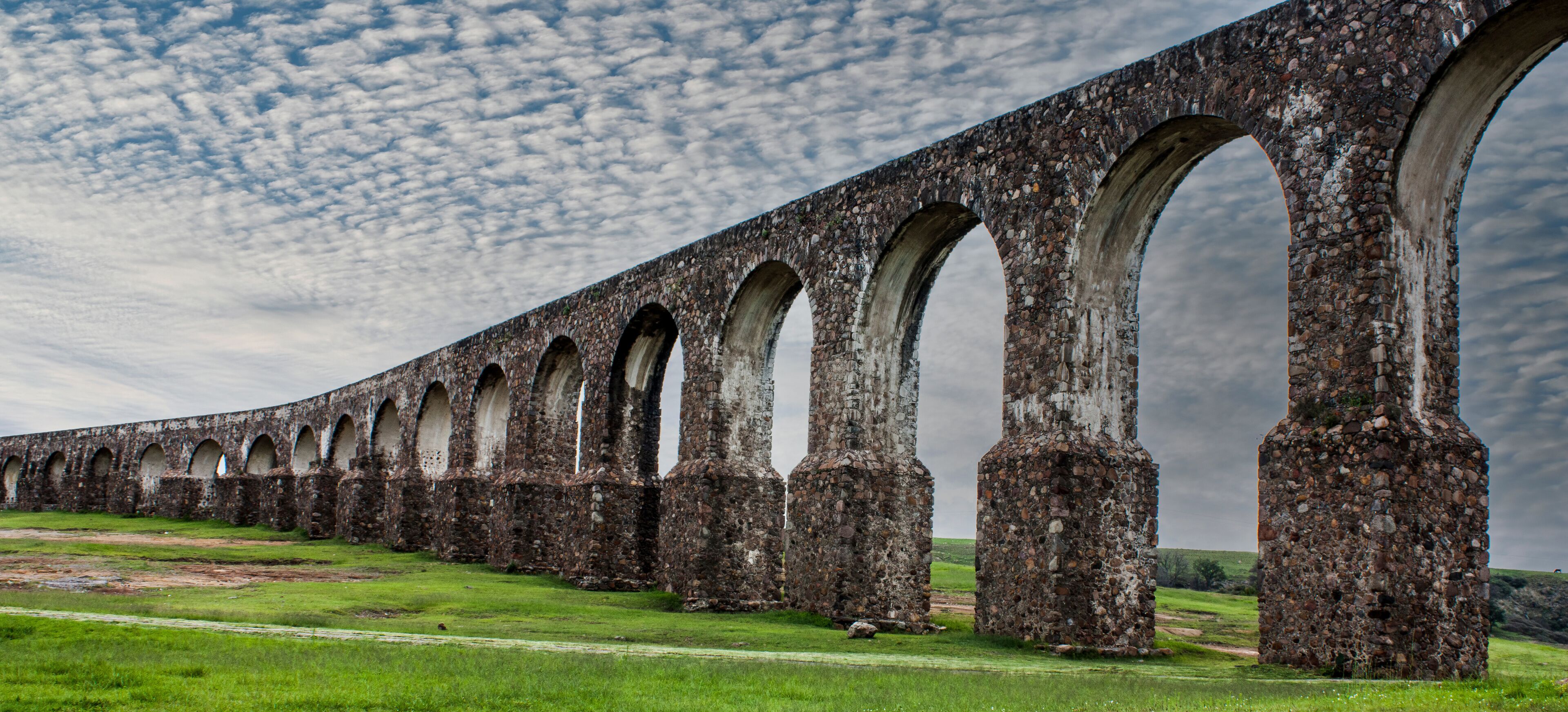 A stone viaduct with multiple arches spanning a grassy field under a cloudy sky.