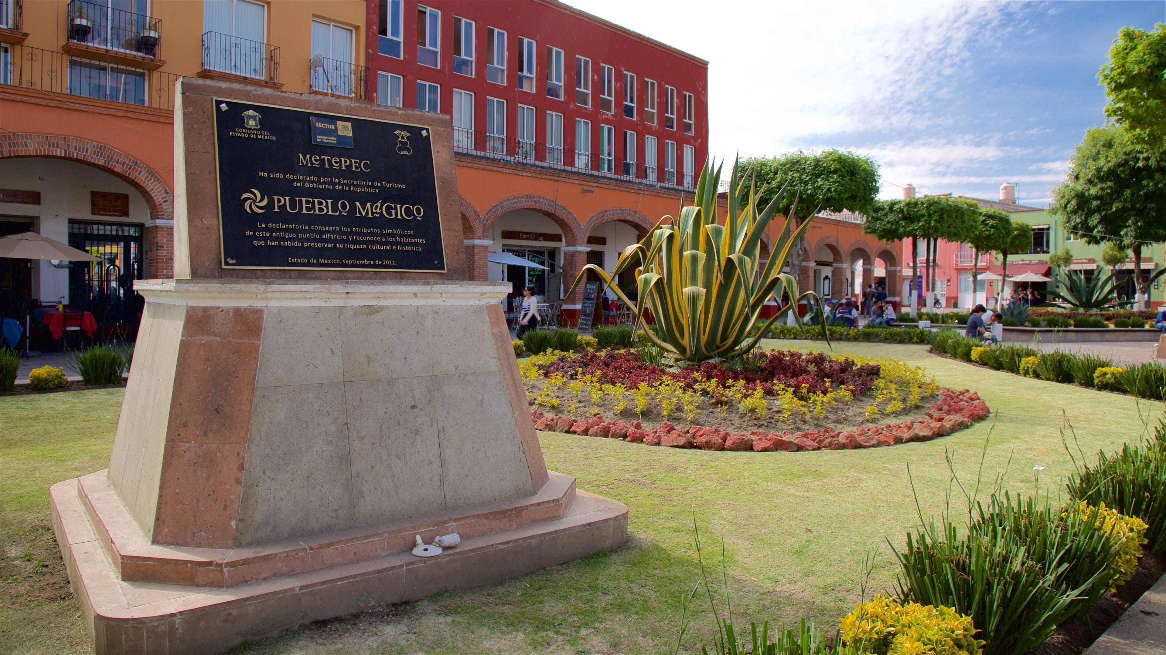 Un monumento con una placa en una plaza pública rodeada por un jardín con diversas plantas y flores.
