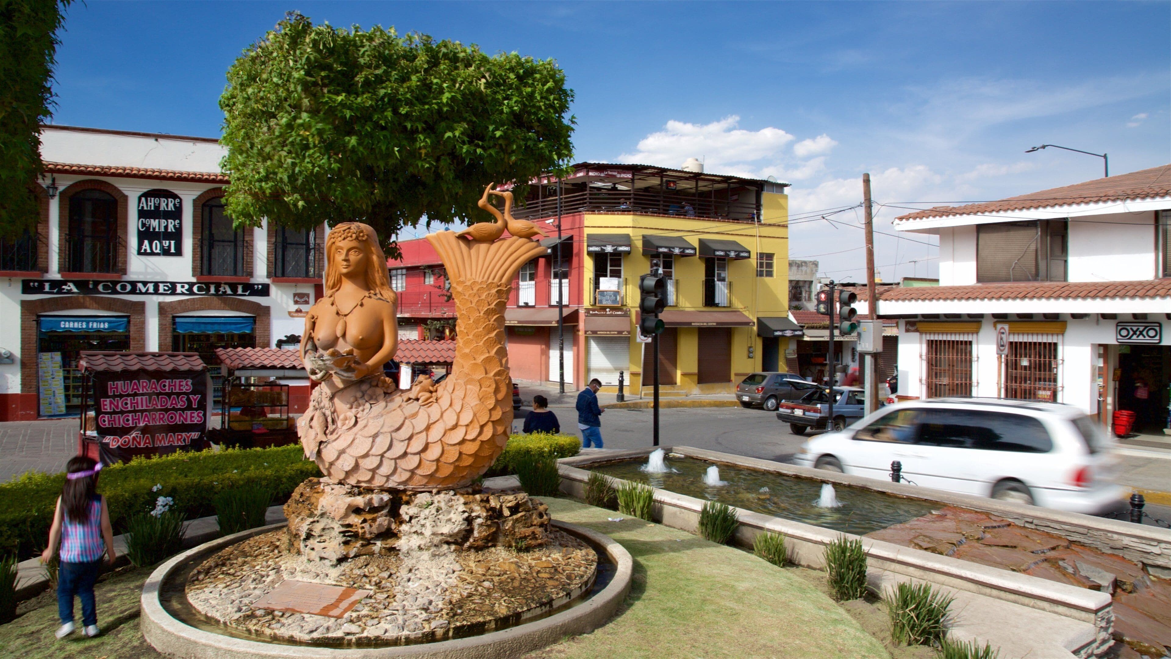 Una gran escultura de una sirena en una fuente, rodeada de un jardín y edificios al fondo.