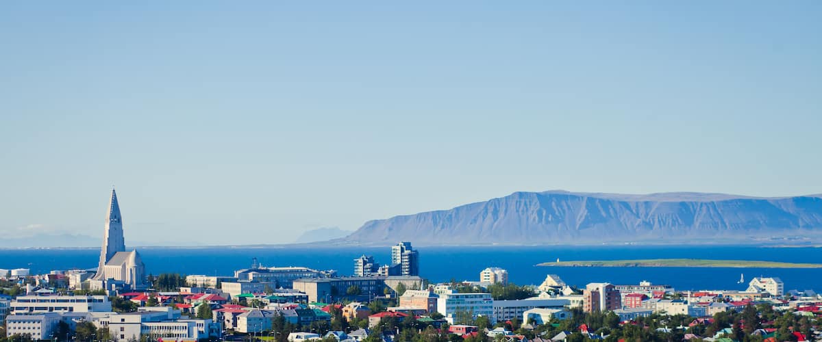 A coastal cityscape with a prominent church spire, a mountain range, and a clear blue sea.