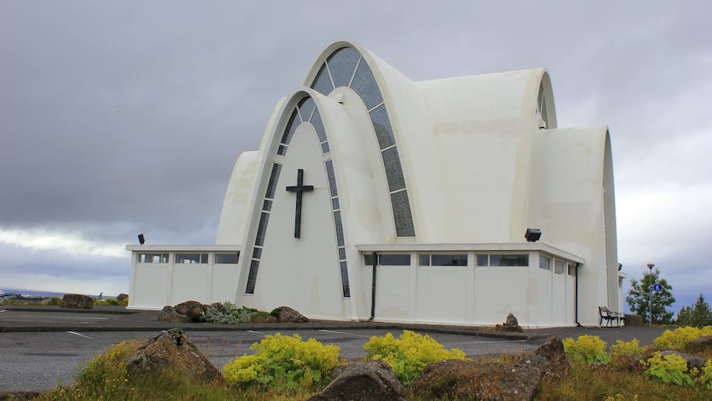 A white church with a cross, large arched windows, and a cloudy sky.