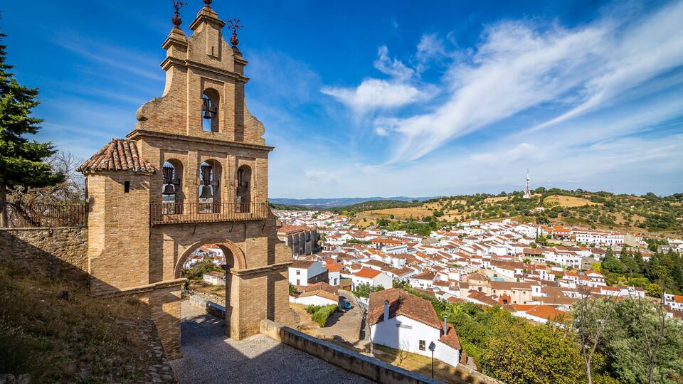 A historic church with a bell tower, surrounded by a town with red-roofed houses and a hilly landscape.