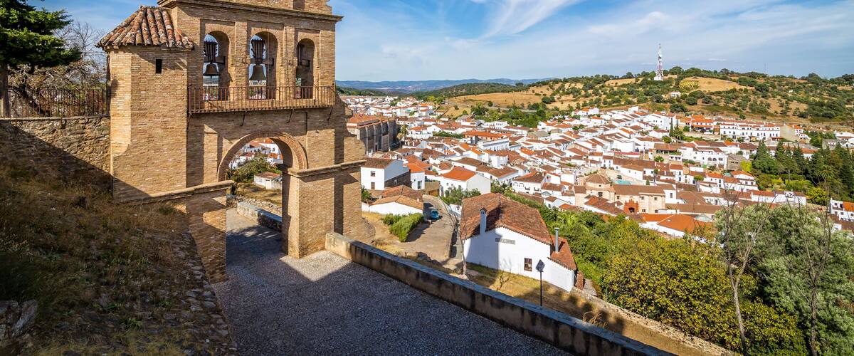 A historic church with a bell tower, surrounded by a town with red-roofed houses and a hilly landscape.