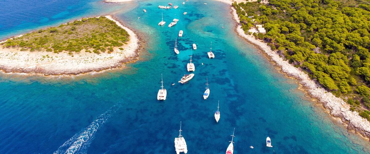 An aerial view of a coastal area with clear turquoise waters, several small islands, and numerous sailboats anchored in the bay.