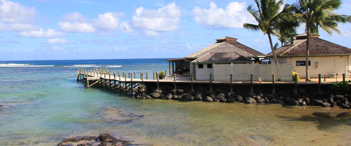 A coastal scene with a wooden pier leading to a white building with a thatched roof, palm trees, and clear blue ocean.