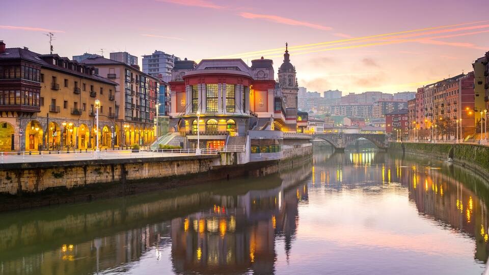 A river with buildings reflecting in the water, a bridge, and a tower in the background.