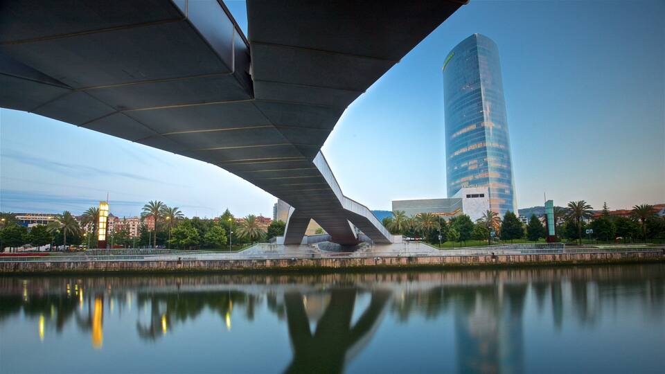 A modern bridge over a calm river with a tall skyscraper in the background.