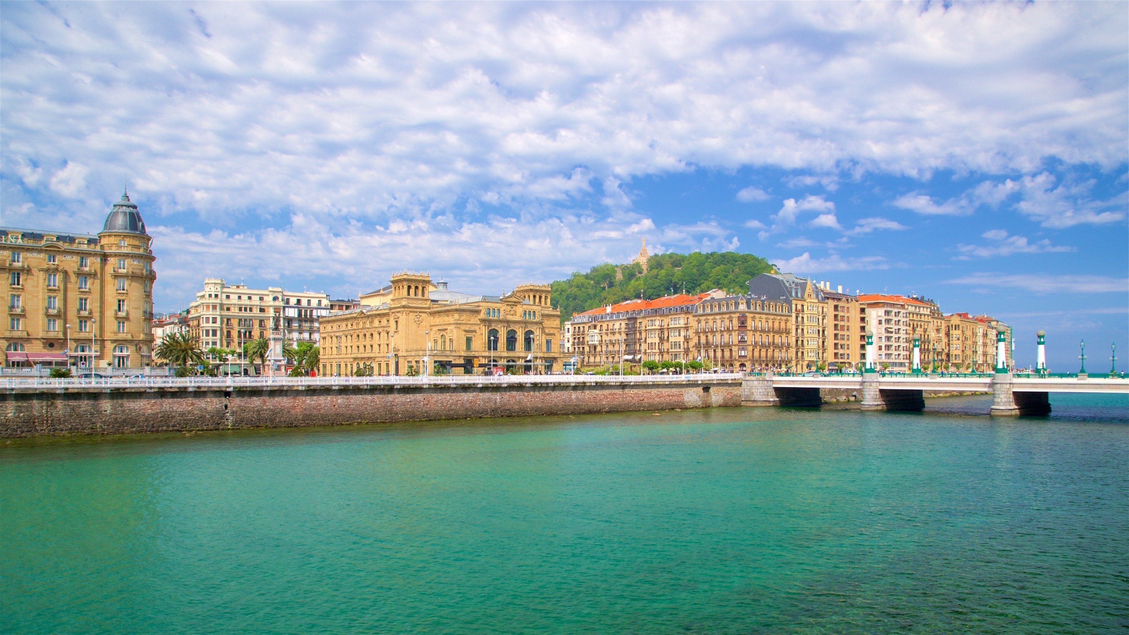 A waterfront cityscape with historic buildings, a bridge, and a hill with a statue.