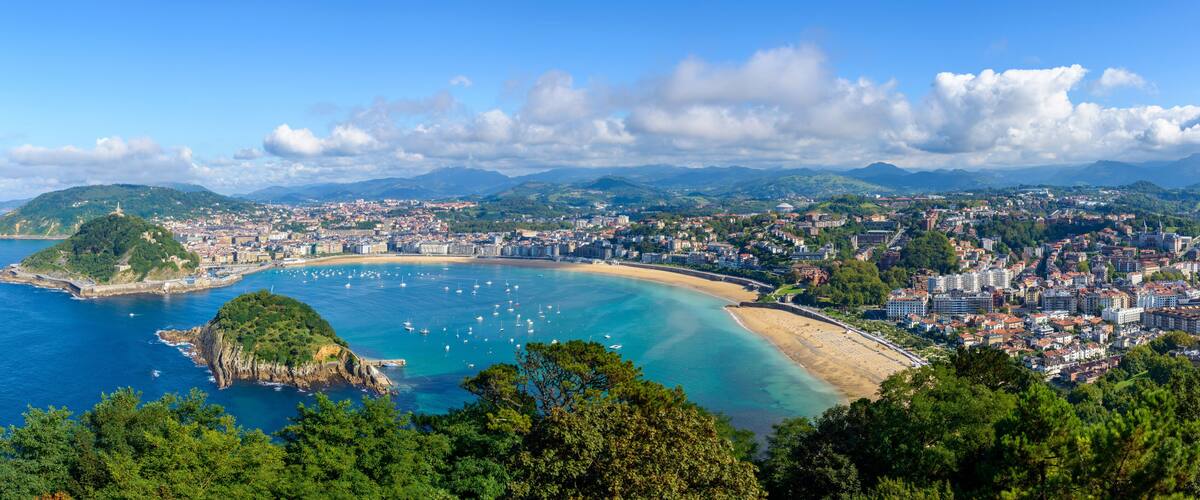 A coastal cityscape with a bay, boats, and a mountainous backdrop.