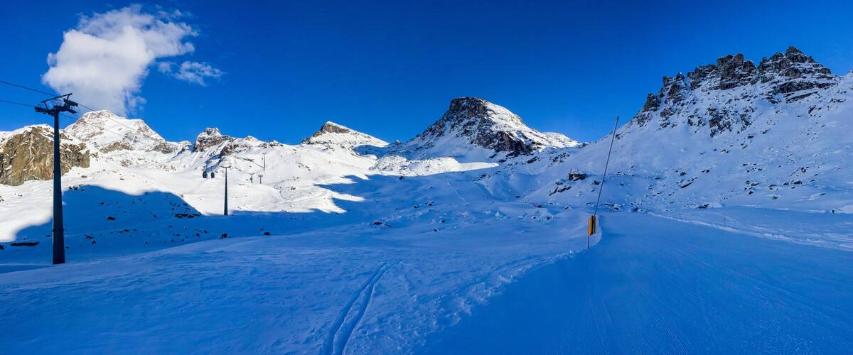 A snow-covered mountain path with ski lifts and rocky peaks under a clear blue sky.
