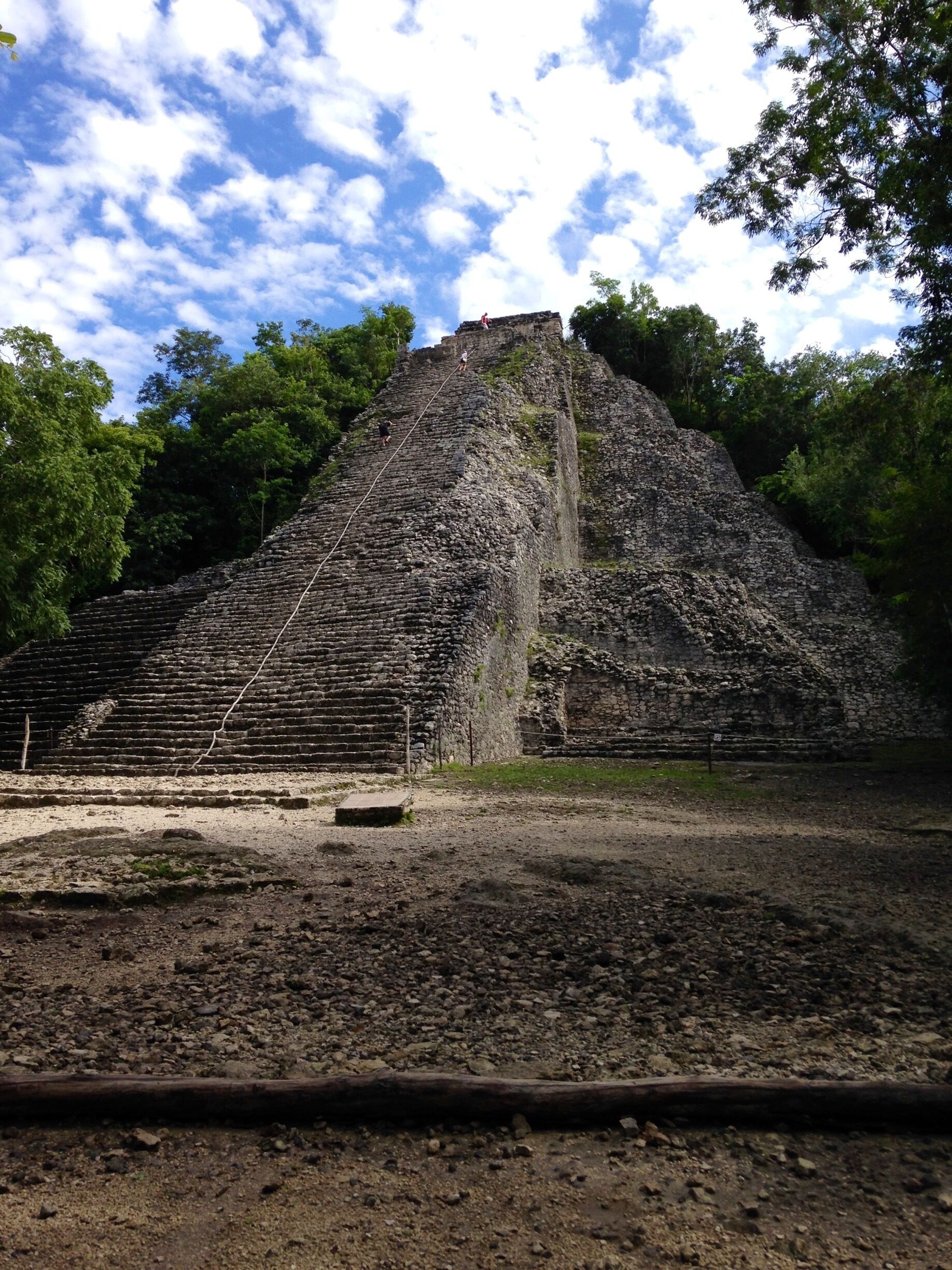A large, ancient stone pyramid with a staircase leading to the top, surrounded by dense green trees.
