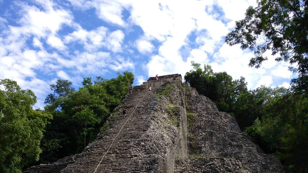 Eine große, uralte SteinsPyramide mit einer Treppe, die zum Gipfel hinaufführt, umgeben von dichtem Grün.