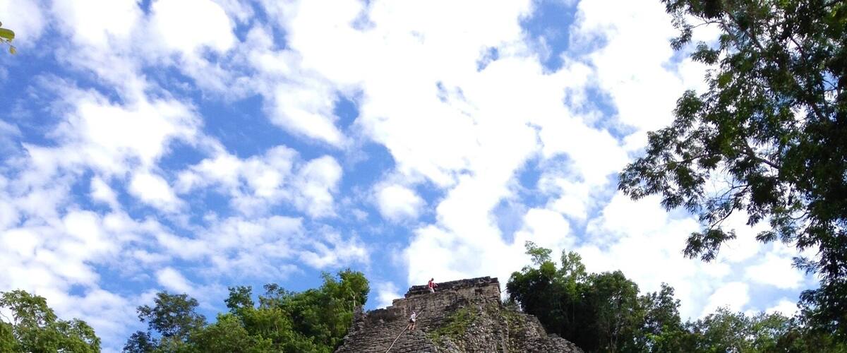Uma grande pirâmide de pedra antiga com uma escadaria que leva ao topo, cercada por árvores verdes densas.