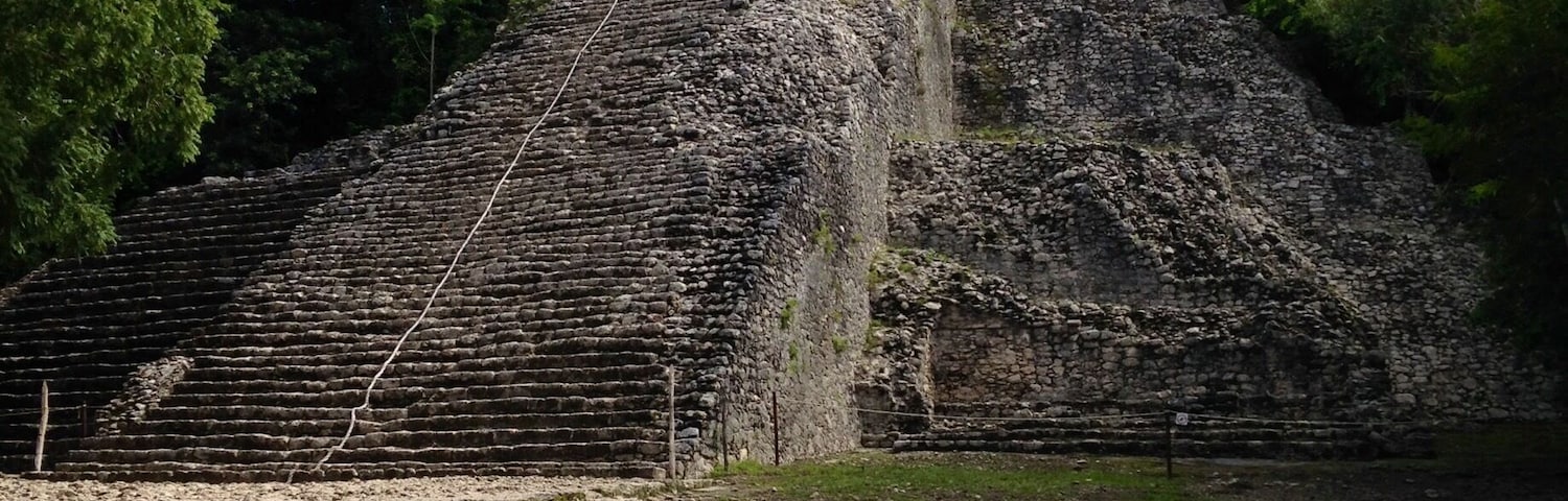 Una gran pirámide de piedra antigua con una escalera que lleva a la cima, rodeada de árboles verdes y frondosos.