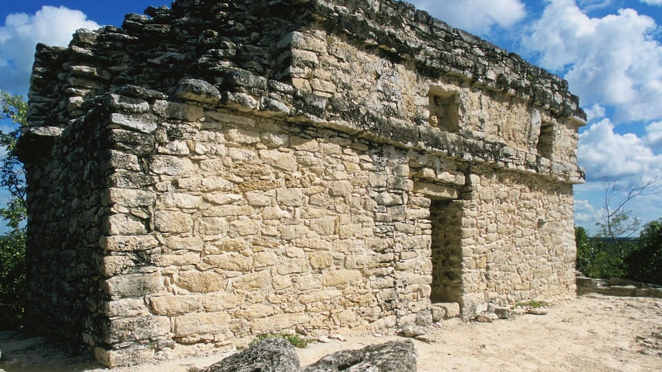 An ancient stone building with a flat roof and a central doorway, surrounded by large rocks and a clear blue sky.