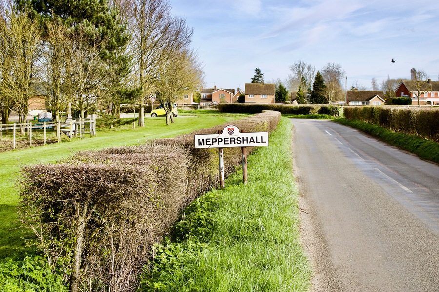 Un camino rural con un cartel 'MEPPERSHALL', setos, árboles y casas al fondo.
