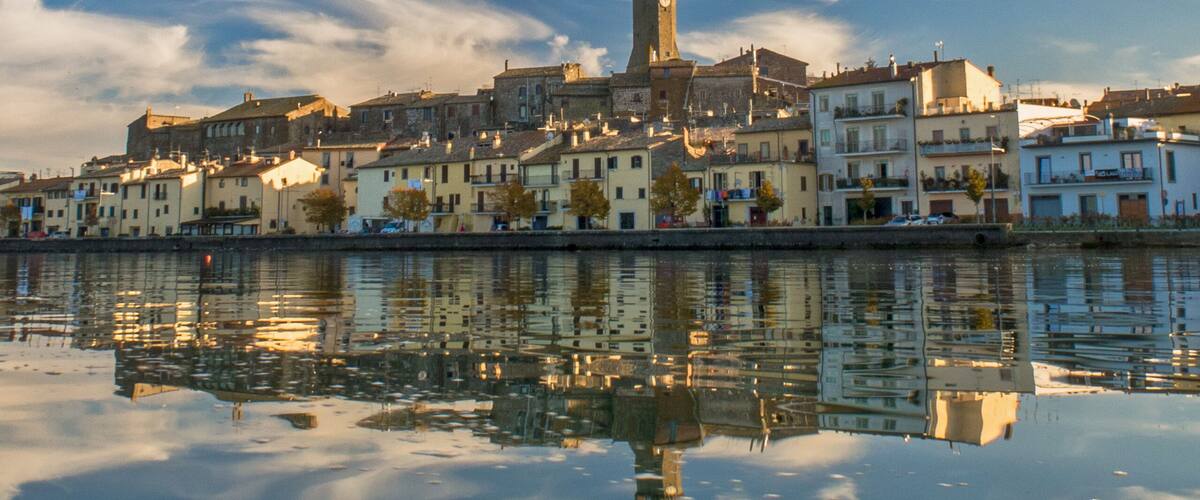 A town with a prominent tower, calm water reflecting the buildings, and a clear blue sky with scattered clouds.