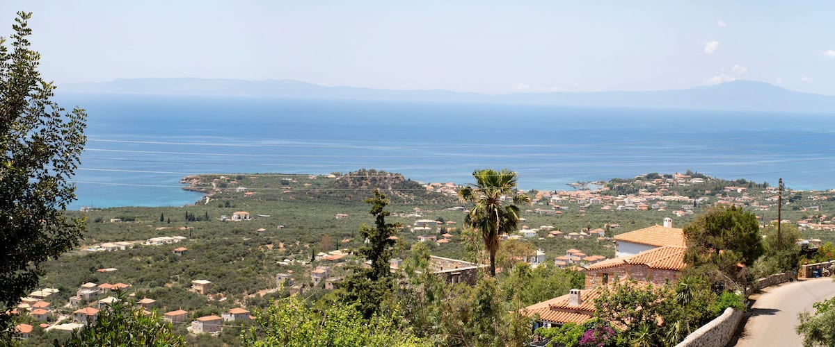 A coastal village with terracotta-roofed houses, a palm tree, and a view of the sea and distant mountains.