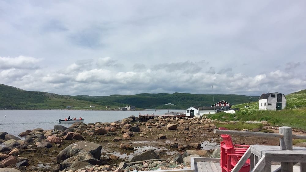 A rocky shoreline with a wooden dock, a small red chair, and a white building in the background.