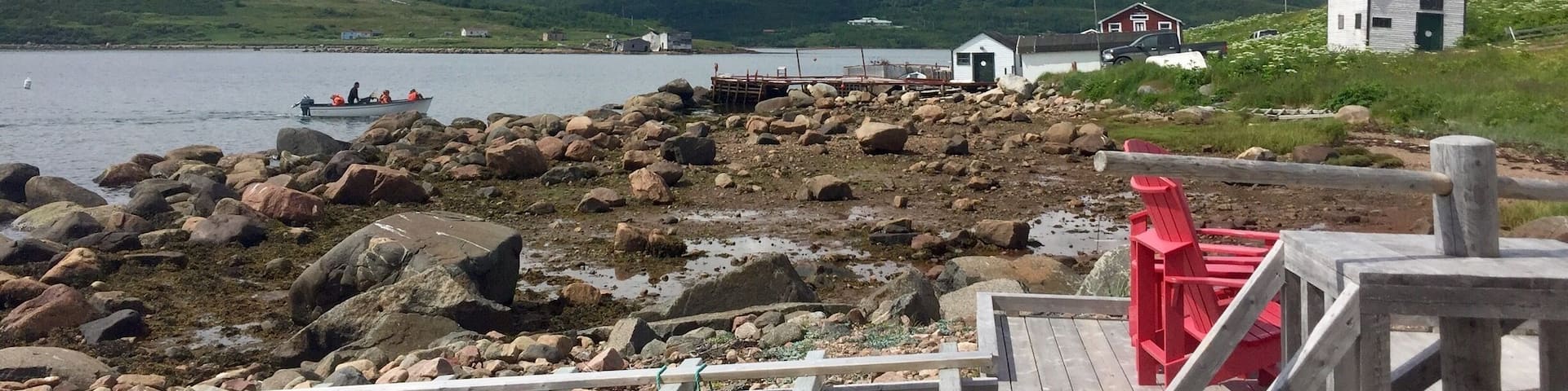 A rocky shoreline with a wooden dock, a small red chair, and a white building in the background.