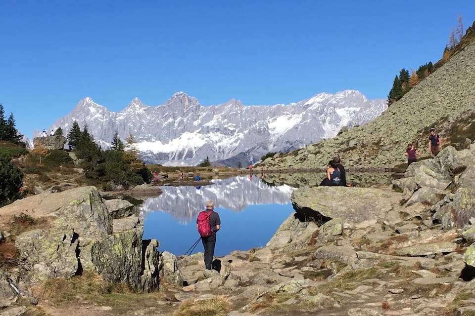 Paisaje montañoso con un lago cristalino que refleja las cumbres, terreno rocoso y excursionistas.