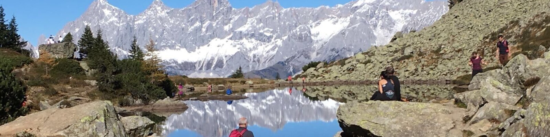 A mountainous landscape with a clear lake reflecting the peaks, rocky terrain, and hikers.