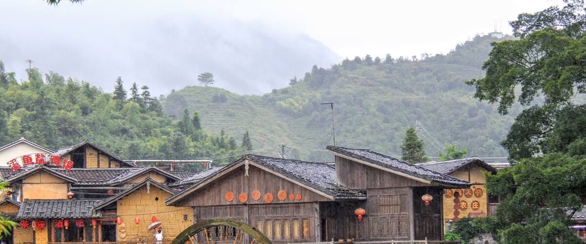 Traditional wooden buildings with a large water wheel, a stone wall, and a mountainous backdrop.