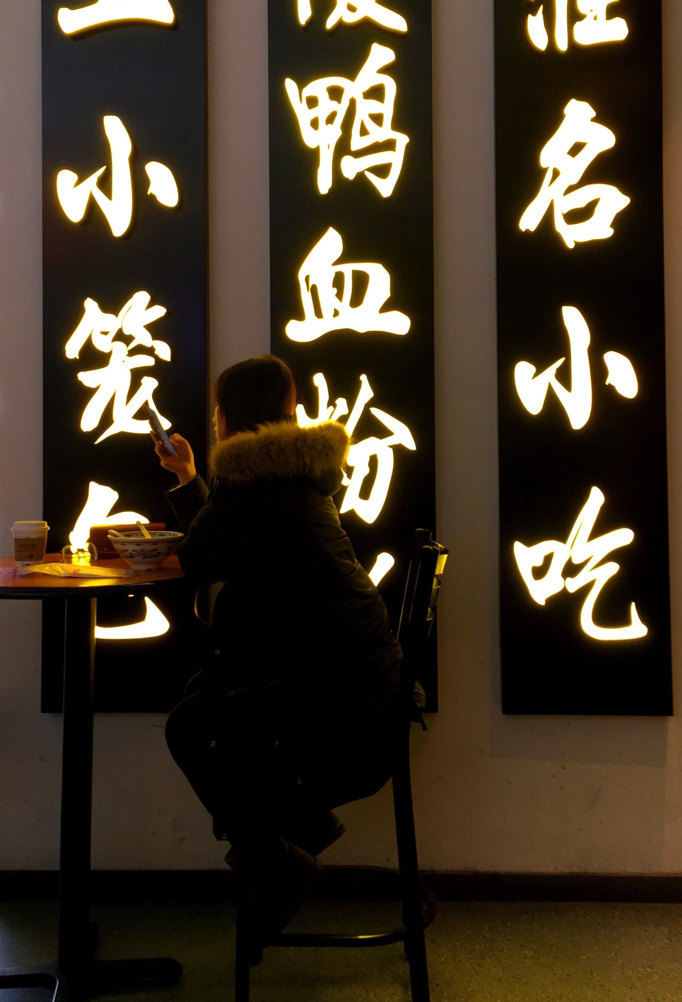 A person sitting at a table with illuminated Chinese characters on the wall behind them.