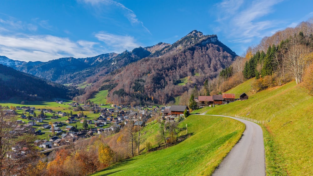 Ein Bergdorf mit einer Serpentinenstraße, Häusern und einem markanten Berggipfel im Hintergrund.