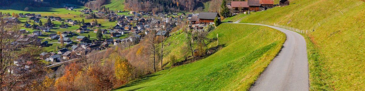 A mountain village with a winding road, houses, and a prominent mountain peak in the background.
