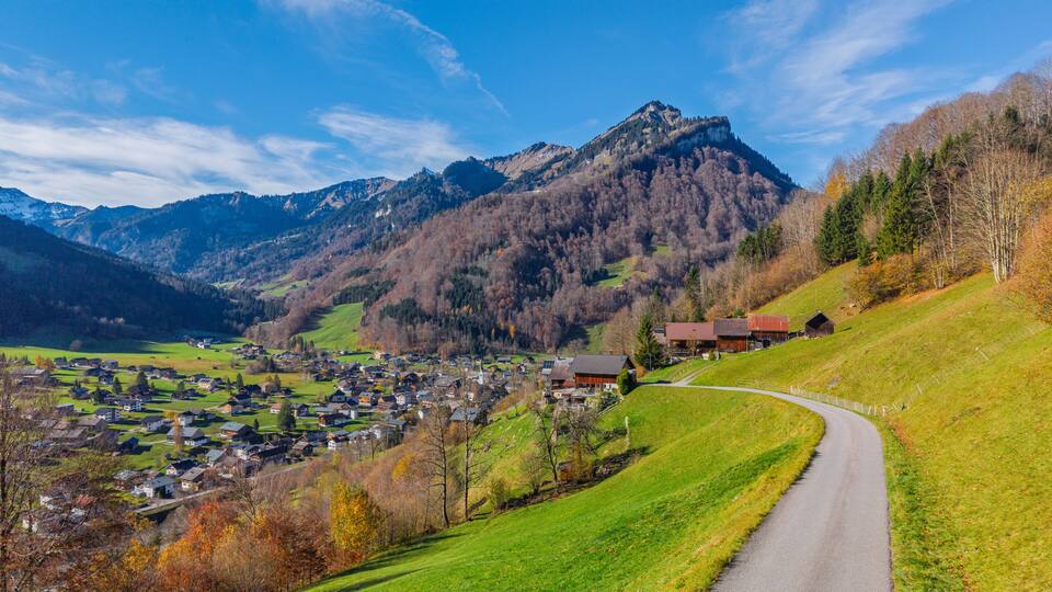 A mountain village with a winding road, houses, and a prominent mountain peak in the background.