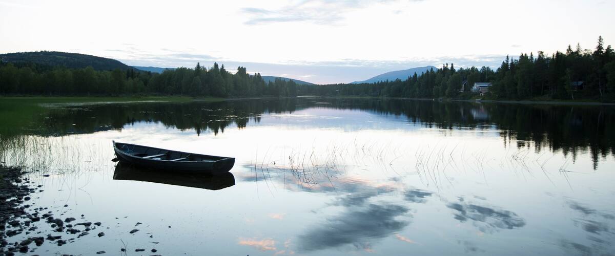 A calm lake with a boat on the left, surrounded by trees and hills in the background.