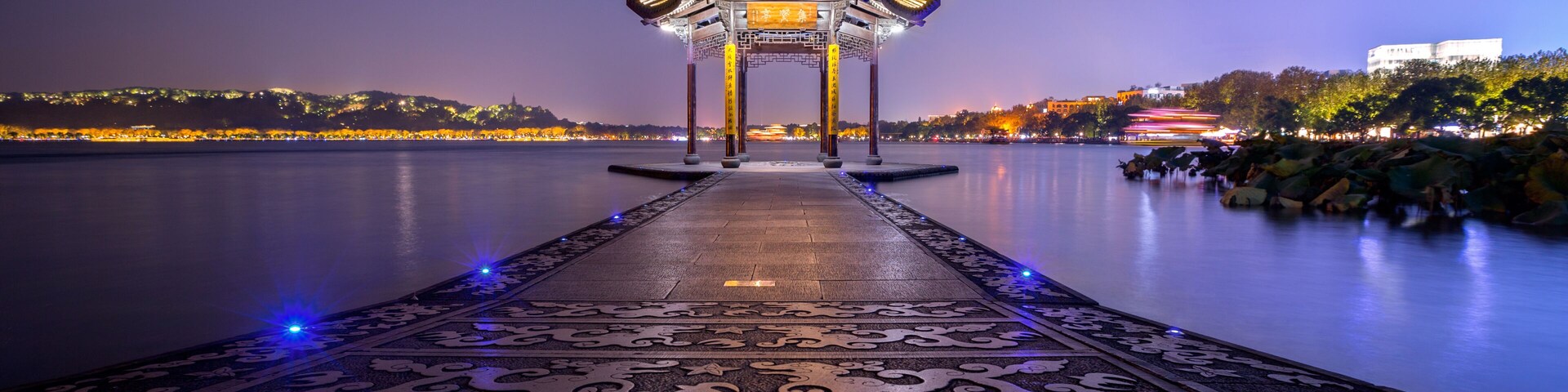 Um pavilhão tradicional com telhado ornamentado e desenhos complexos no chão, com vista para um corpo d'água calmo.