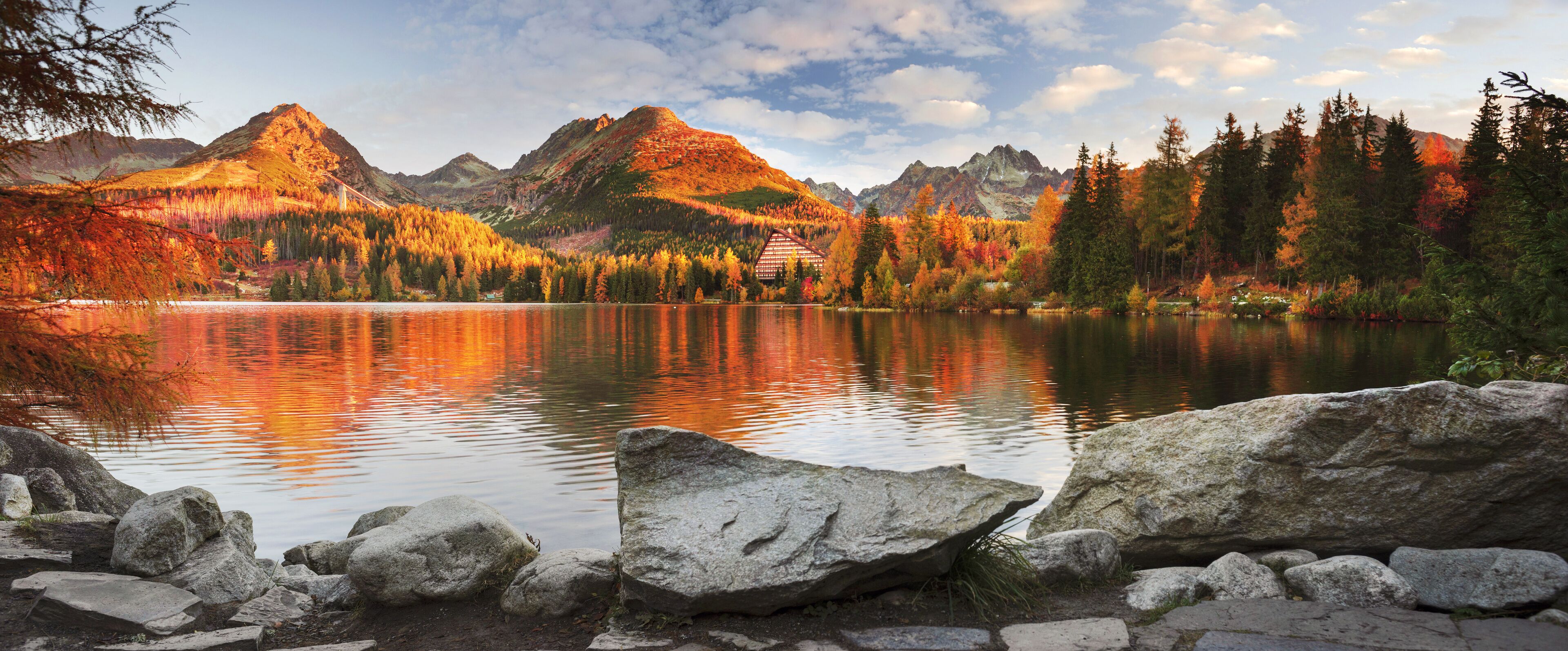 Un lago de montaña rodeado de follaje otoñal, una cabaña y un cielo despejado.