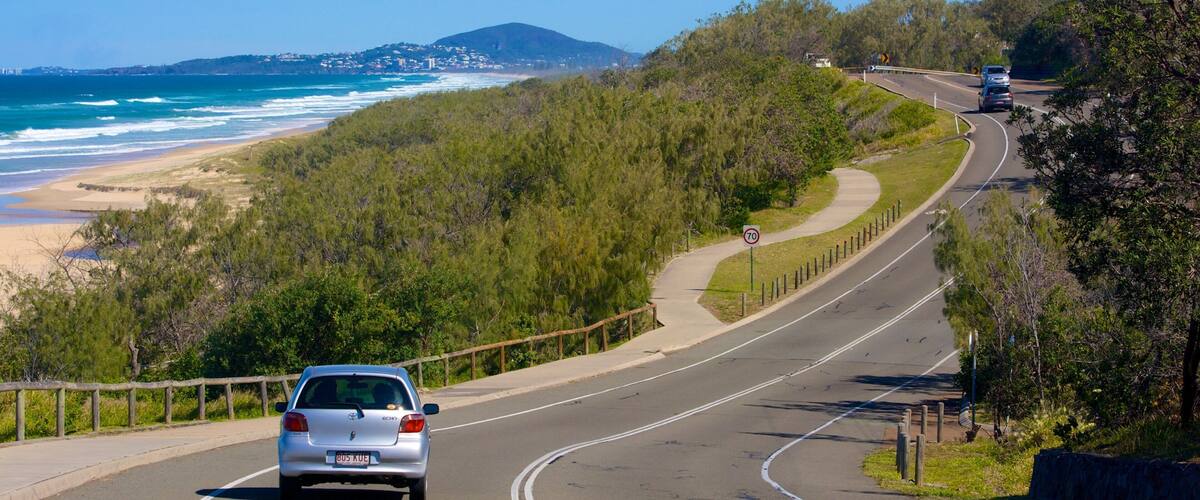 A coastal road with a beach, forested hills, and a clear blue sky.