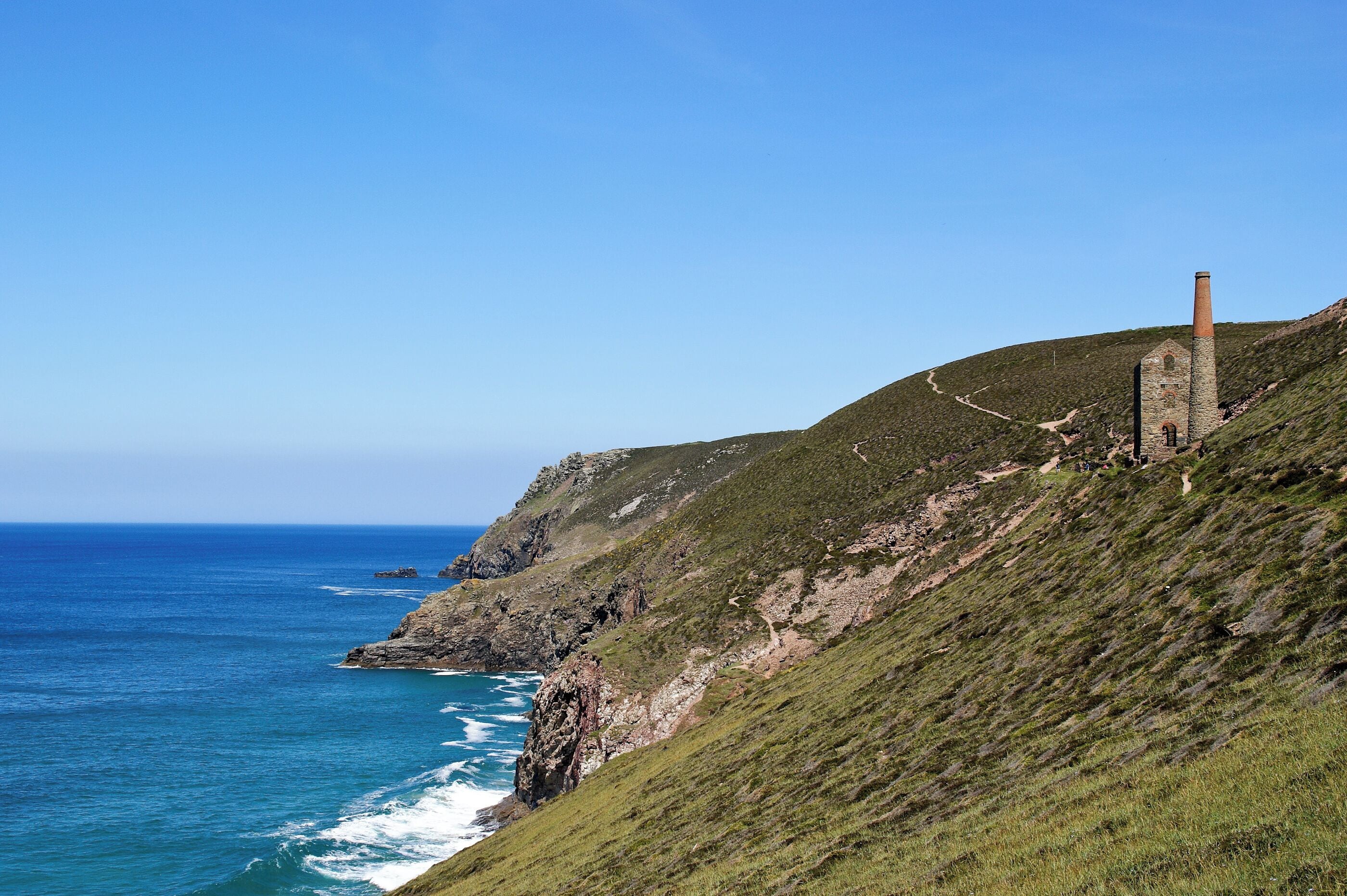 A coastal landscape with a grassy hill, a rocky coastline, and a tall chimney-like structure.