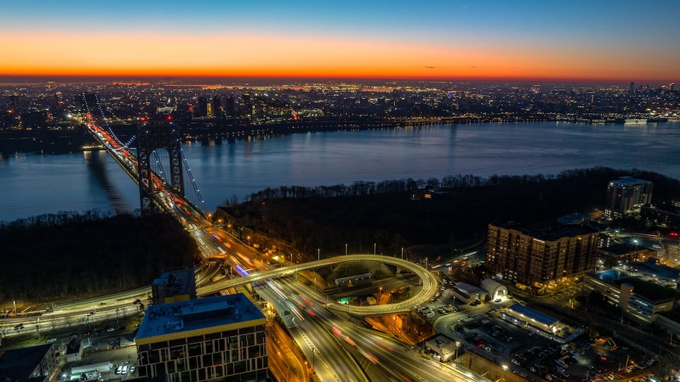 A cityscape at dusk with a prominent bridge, a river, and a highway interchange.