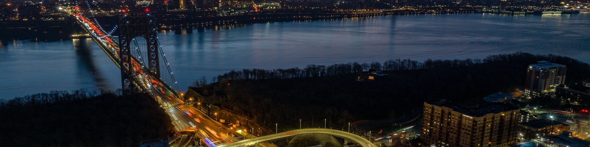 A cityscape at dusk with a prominent bridge, a river, and a highway interchange.