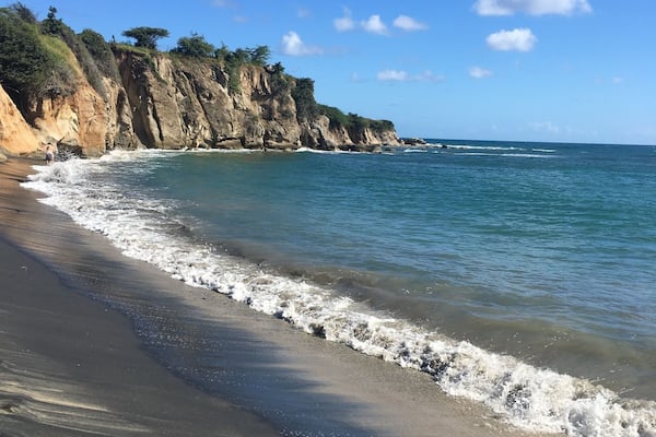 A sandy beach with gentle waves, a rocky cliff, and a clear blue sky.