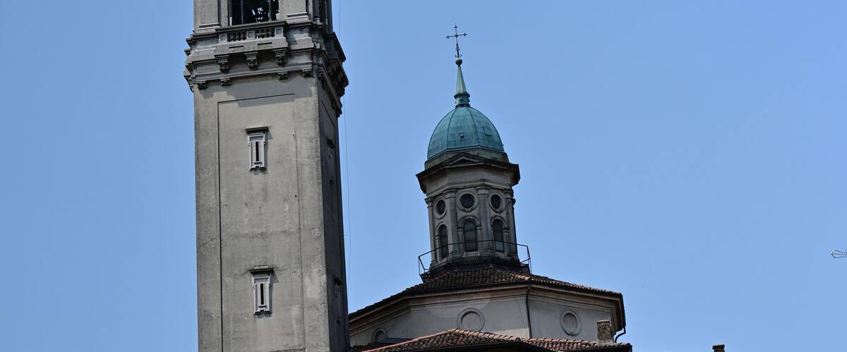 A tall clock tower with a dome and a smaller spire, surrounded by buildings and greenery.