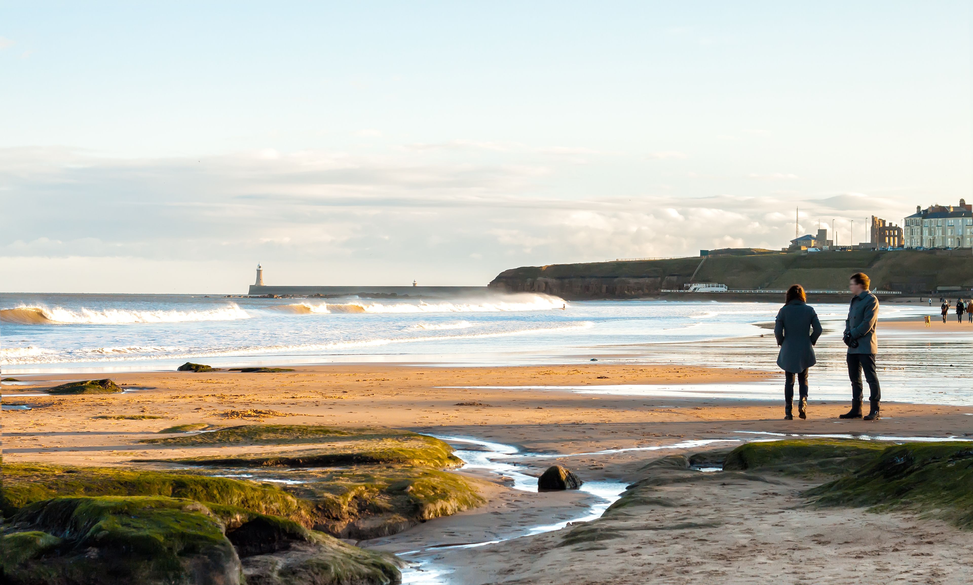 A beach with waves, a lighthouse, and buildings in the background.