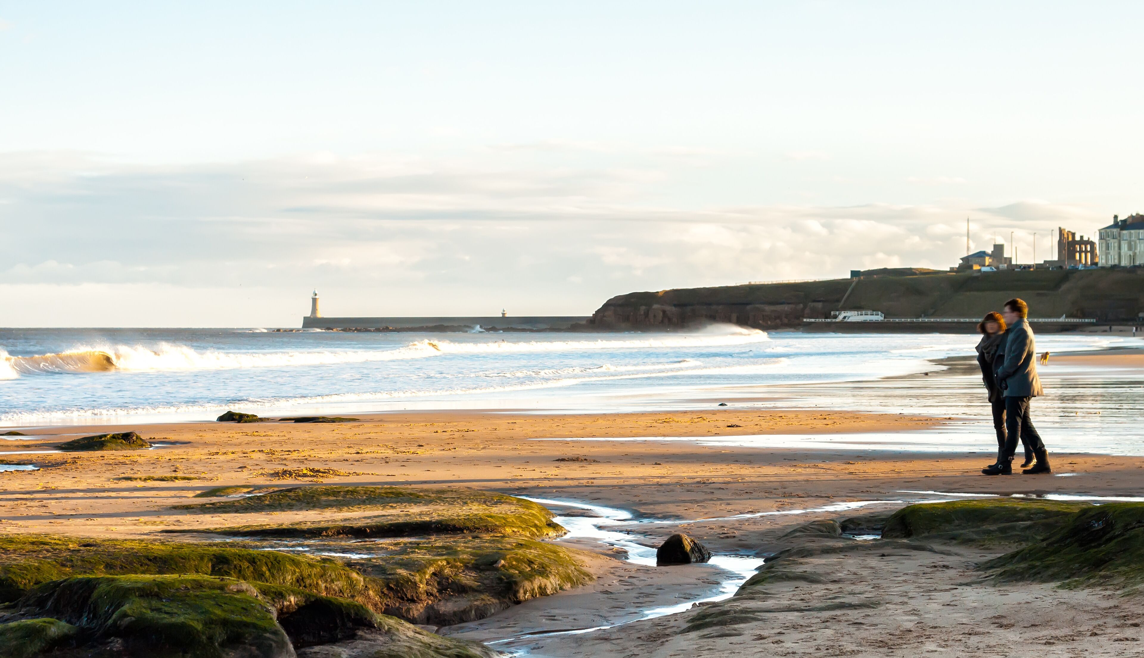 A beach with waves, a lighthouse, and a coastal town in the background.