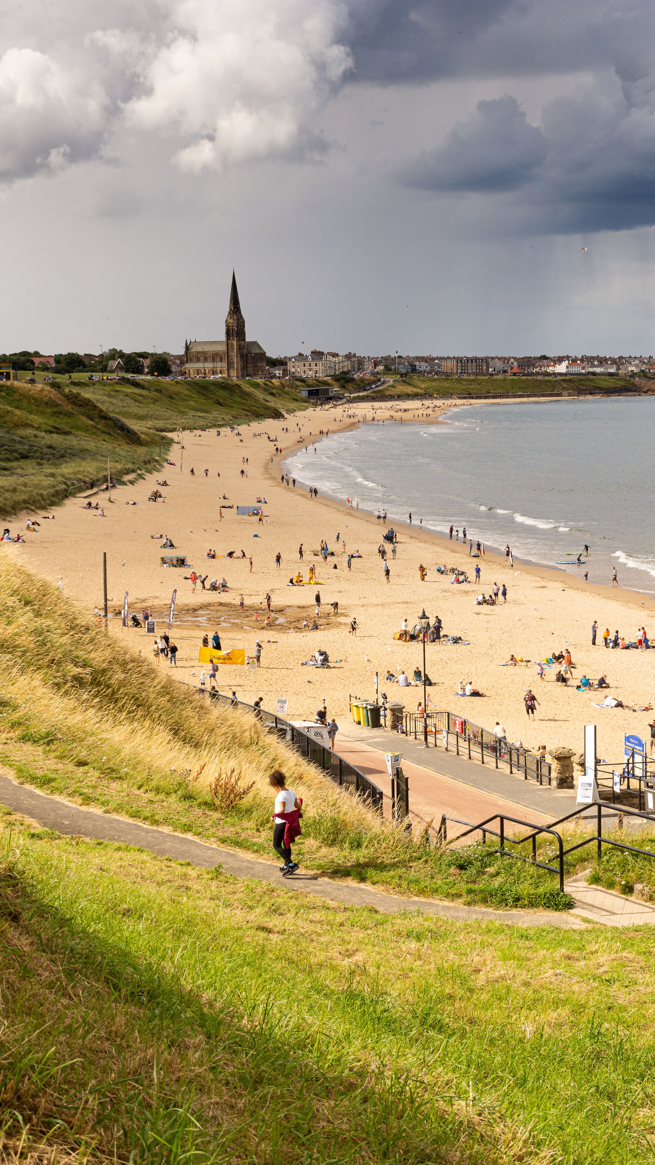 A coastal beach with people sunbathing and walking, a church in the background, and a grassy hill in the foreground.