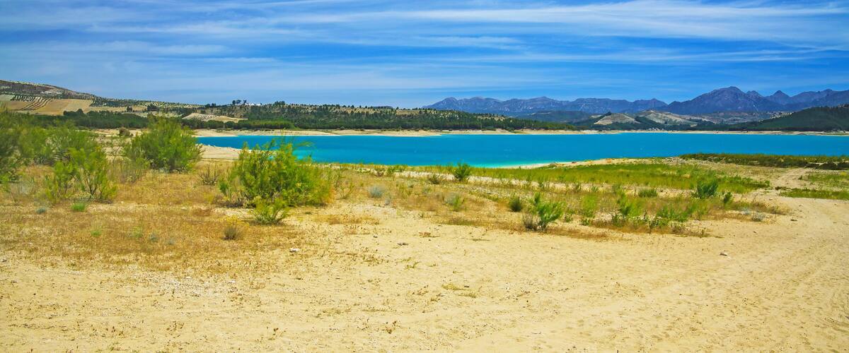 A coastal landscape with a clear blue sea, sandy beach, and distant mountains.