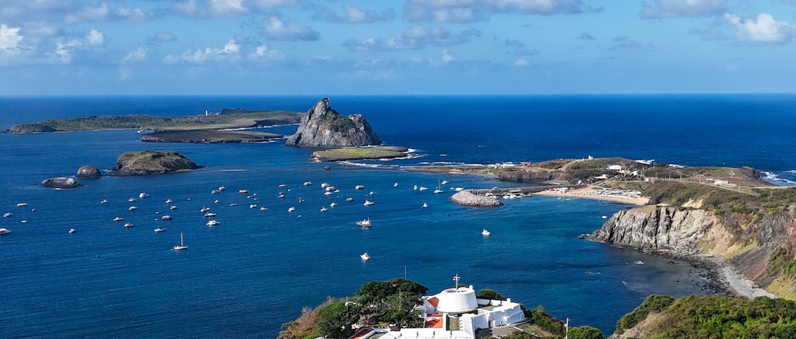 Paisaje costero con un edificio blanco en un acantilado, una playa y barcos en el agua.