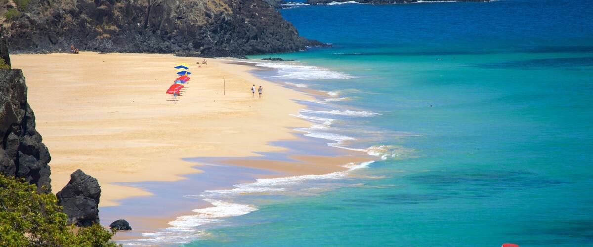 A beach with clear turquoise waters, a sandy shore, and rocky outcrops.