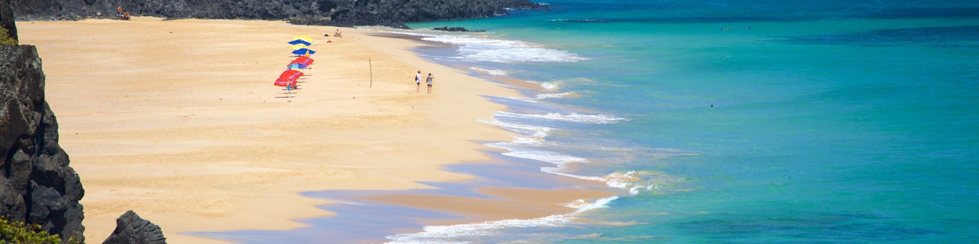 A beach with clear turquoise waters, a sandy shore, and rocky outcrops.